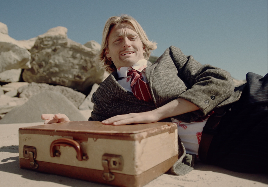 Josiah Amico laying on beach in formal suit with with a briefcase