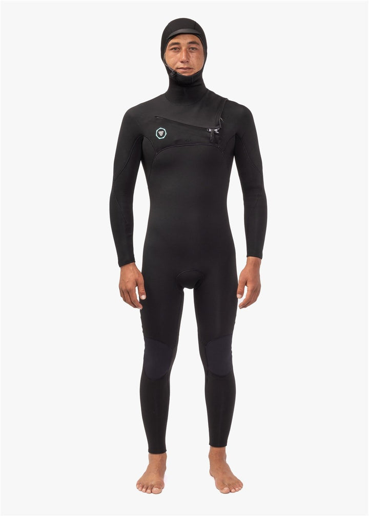 A person wearing a full hooded chest zip black wetsuit, standing against a white background.